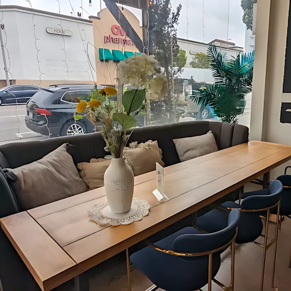 A long wooden table with a vase of flowers and blue bar stools in a cafe by a window.