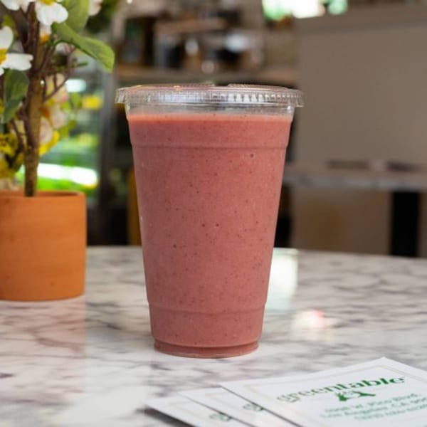 A Green Table Cafe strawberry smoothie in a clear cup on a marble table with a plant.