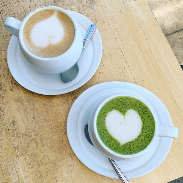 Two heart-shaped lattes, one coffee and one matcha, on a light wooden table.