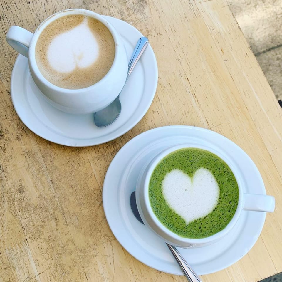 Two heart-shaped lattes, one coffee and one matcha, on a light wooden table.
