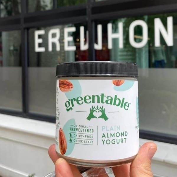 Hand holding a Greentable almond yogurt jar in front of a window with "EREWHON" visible.