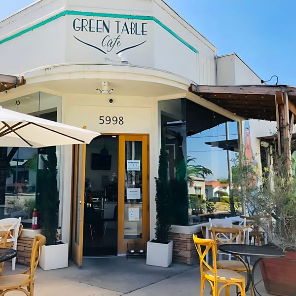 The Green Table Cafe exterior with outdoor seating, a white umbrella, and a wooden door.