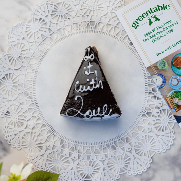 A slice of chocolate cake with "Do it with Love" written in white icing, next to a Green Table Cafe card.