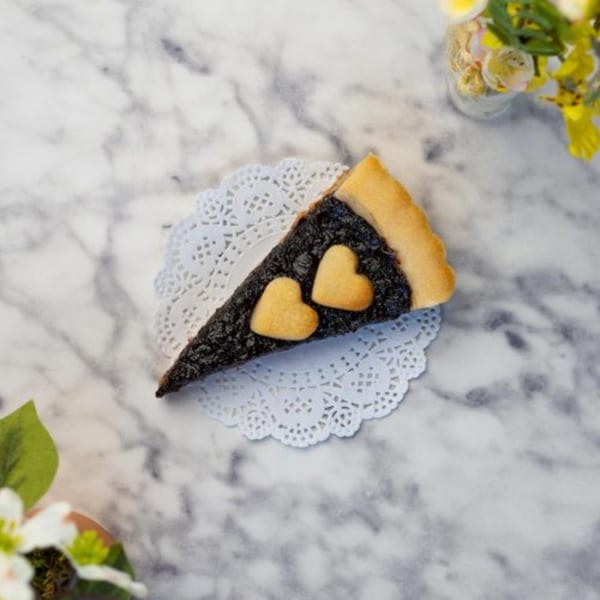 A slice of dark berry tart with two heart-shaped cookies on a white doily.