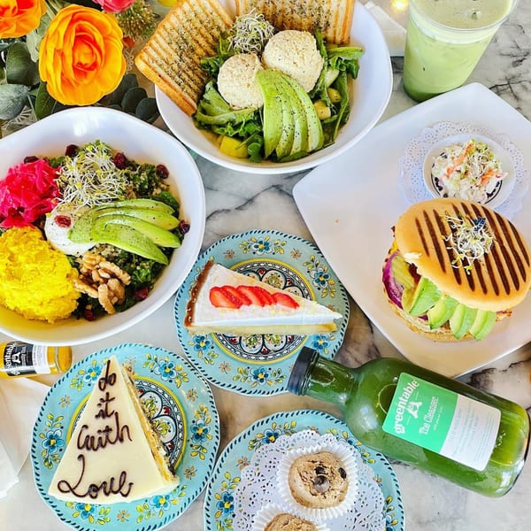 A Green Table Cafe spread of healthy bowls, a panini, desserts, and green drinks on a marble table.