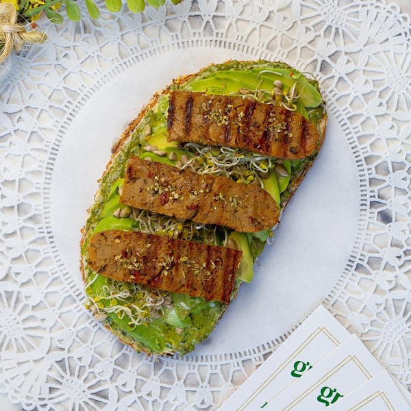 Avocado toast with grilled plant-based protein, sprouts, and seeds on a white lace placemat.