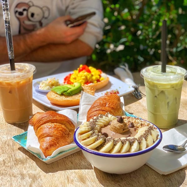 Outdoor cafe table with iced coffee, matcha, croissants, a breakfast bagel, and a smoothie bowl.