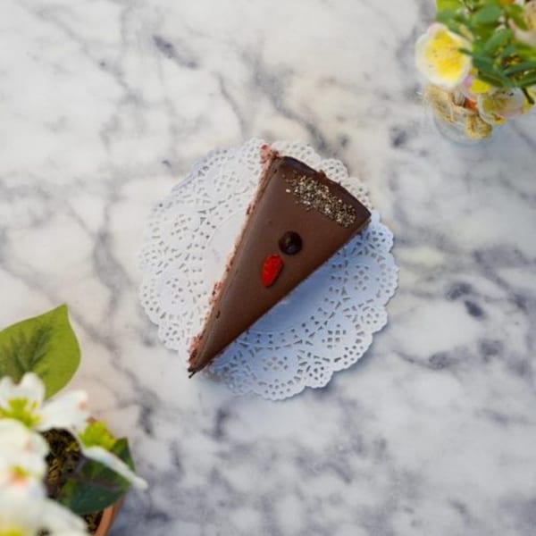 A slice of chocolate cake with sprinkles and a red candy on a white doily, on a marble table.