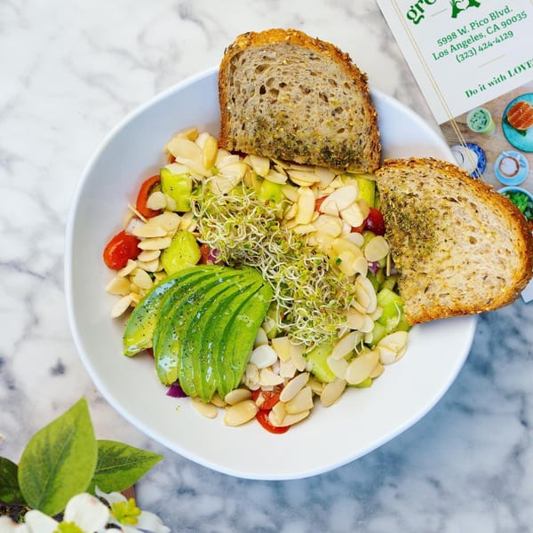 A vibrant Green Table Cafe salad with avocado, sprouts, almonds, and cherry tomatoes, served with two slices of bread.