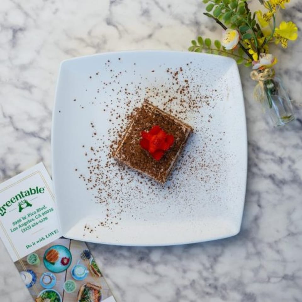 A Green Table Cafe tiramisu dessert with cocoa powder and a red flower on a white plate.