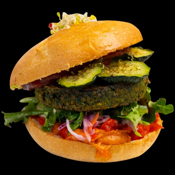 A Green Table Cafe vegan burger with a dark patty, zucchini, and fresh vegetables on a bun.