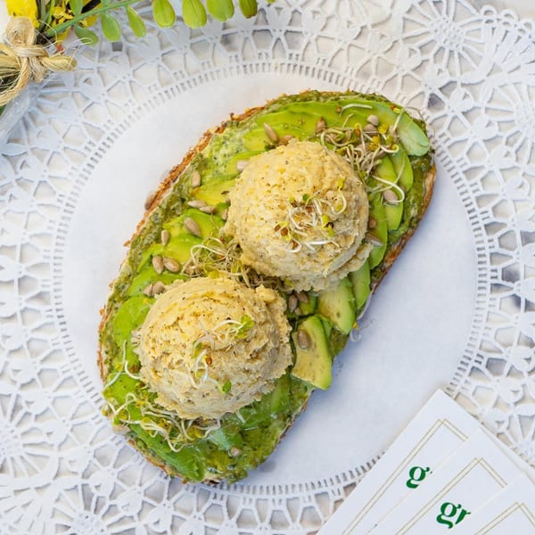 Avocado toast with two scoops of hummus, sunflower seeds, and sprouts on a white lace placemat.