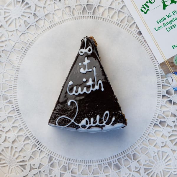 A slice of chocolate cake with white icing spelling "do it with love" on a doily.