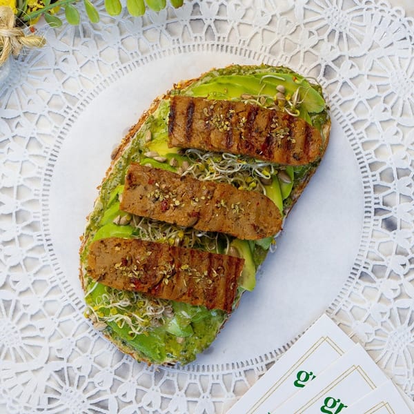 Avocado toast with grilled tempeh, sprouts, and seeds on a white lace placemat.